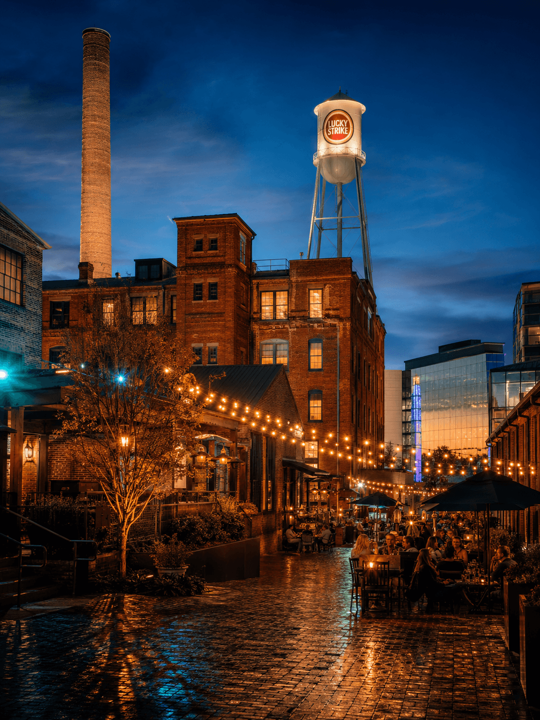 The Lucky Strike water tower above the American Tobacco Campus at dusk, with string lights over a courtyard of restaurant tables.