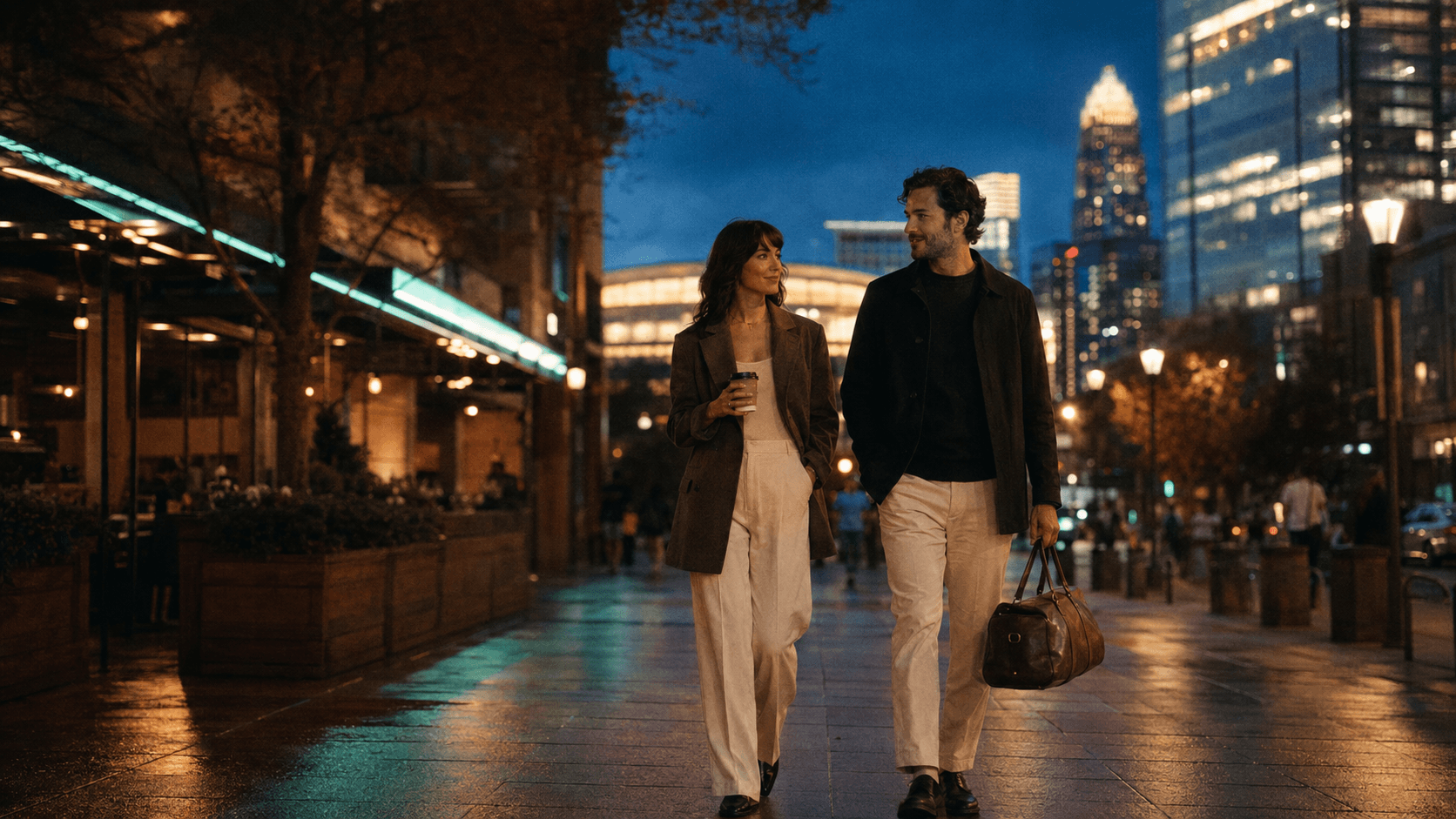 A couple walking through Uptown Charlotte at dusk, the Bank of America crown skyline glowing behind them on a rain-slick street.